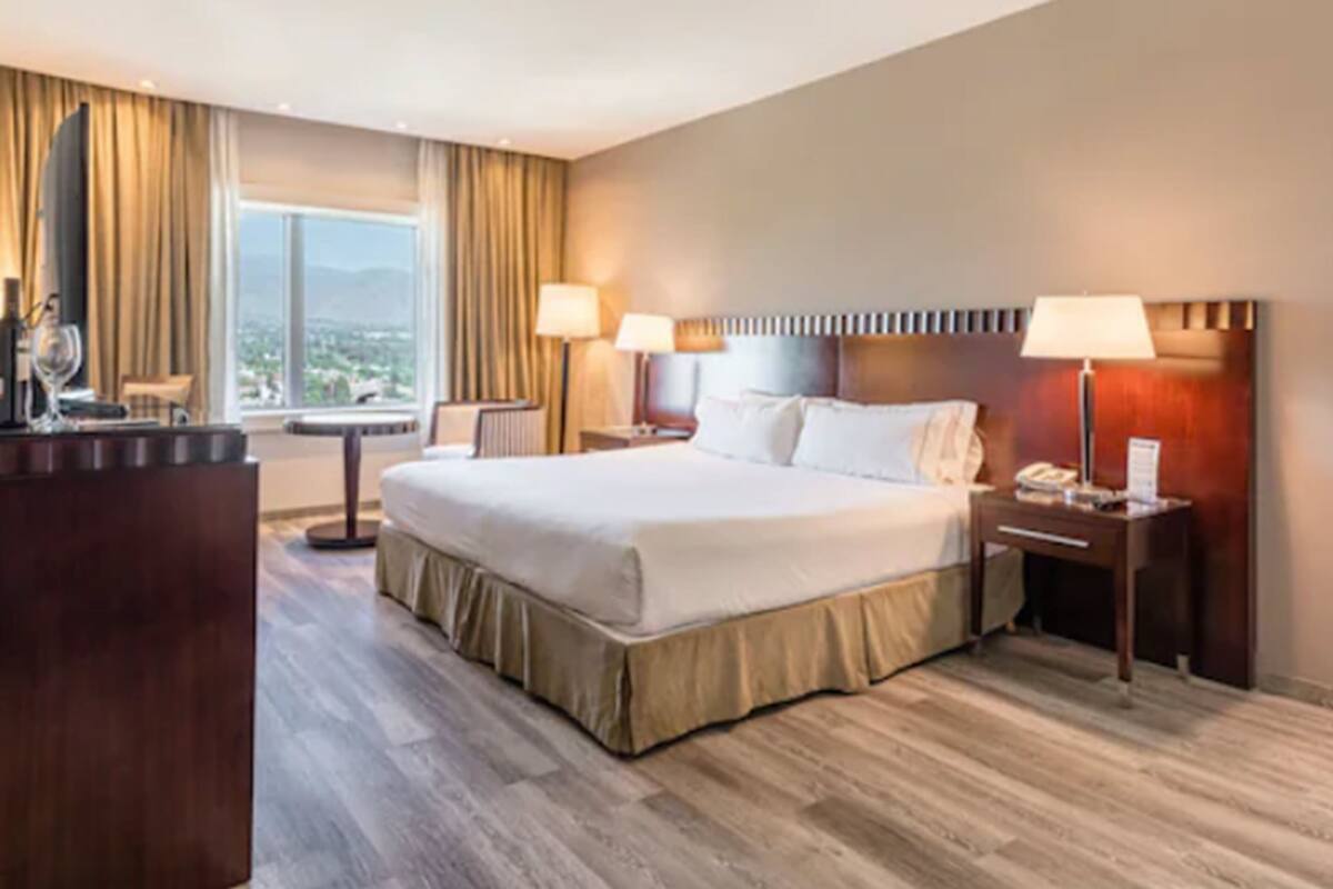 A hotel room with a large bed, two bedside tables with lamps, a desk with a chair, and a view of the city through large windows.