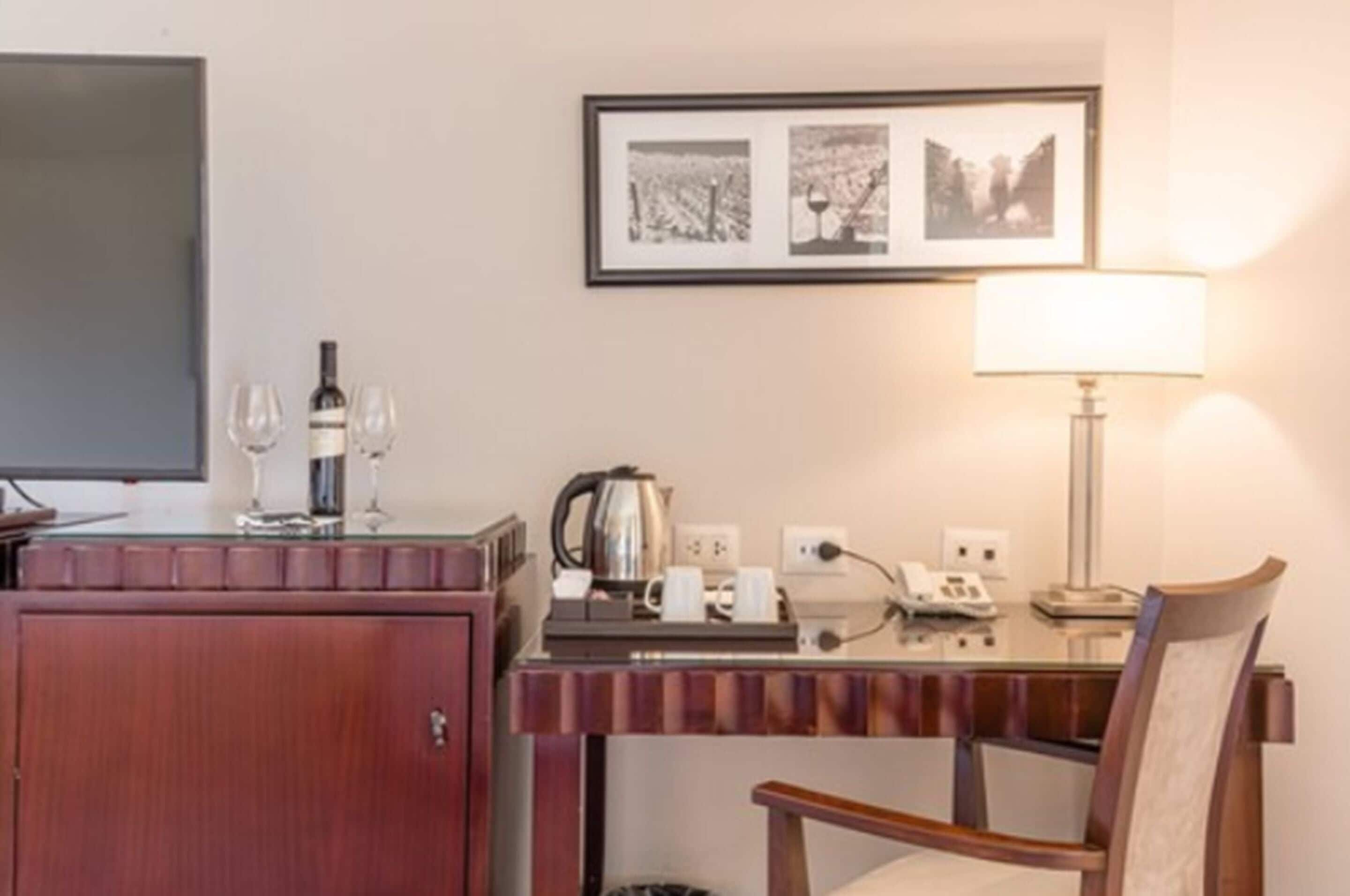 Habitación de hotel con un mueble de madera, un escritorio con hervidor, una lámpara y una silla. En la pared hay cuadros enmarcados y un televisor a la izquierda.