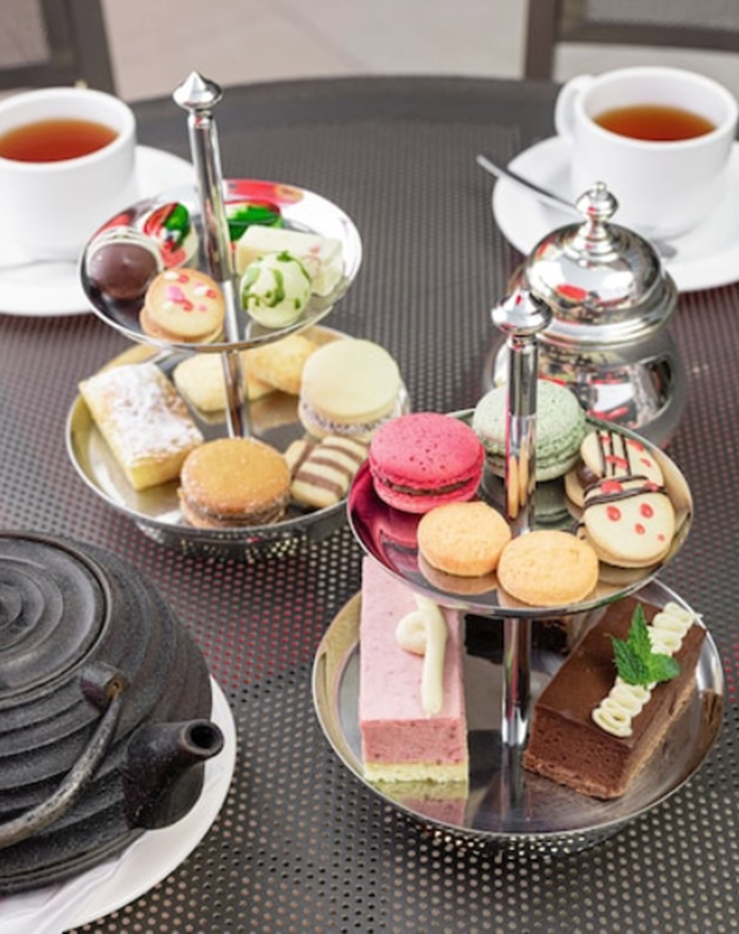 Una bandeja escalonada con varios pasteles y postres, un hervidor de té y dos tazas de té sobre una mesa.
