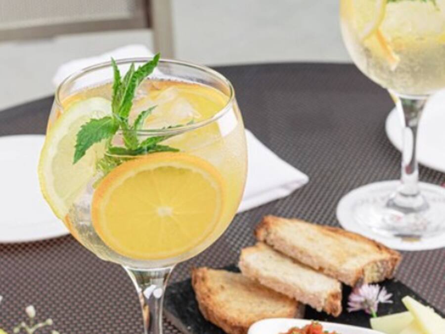 A table setting with two glasses of a citrus beverage, slices of orange, mint leaves, and a small bowl of tomato-based condiment. There are also slices of cheese and bread on the table.