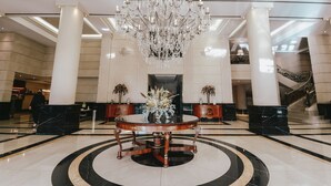 A grand lobby with a large chandelier, a round table with a vase, and a staircase in the background. - Diplomatic Hotel (Mendoza)