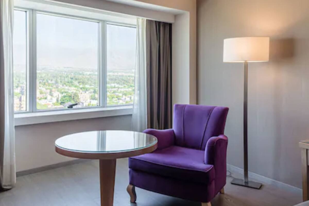 A room with a purple armchair, a round glass table, a floor lamp, and a large window with a view of a cityscape.