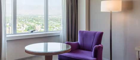Una habitación con un sillón púrpura, una mesa redonda de vidrio, una lámpara de pie y un amplio ventanal con vistas a un paisaje urbano.