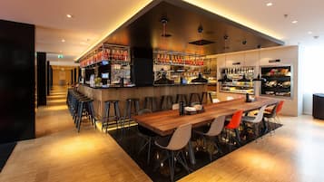 A modern bar area with a long wooden table and several stools, a bar counter with a variety of bottles, and a seating area with a mix of chairs.