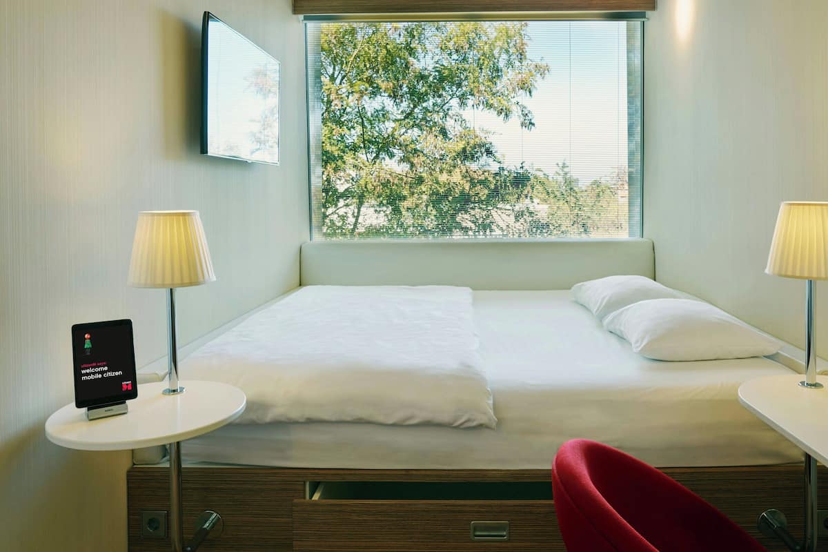 Een hotelkamer met een bed, een tafeltje met een lamp, een rode stoel en een televisie die aan de muur is bevestigd.