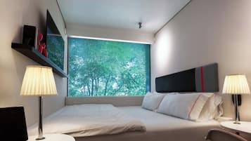 A modern hotel room with a bed, two bedside tables, a TV, and a window with a view of trees.