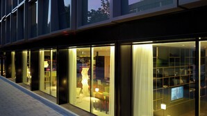 A modern hotel exterior with large glass windows, a brick-paved walkway, and a visible interior with seating and a TV.