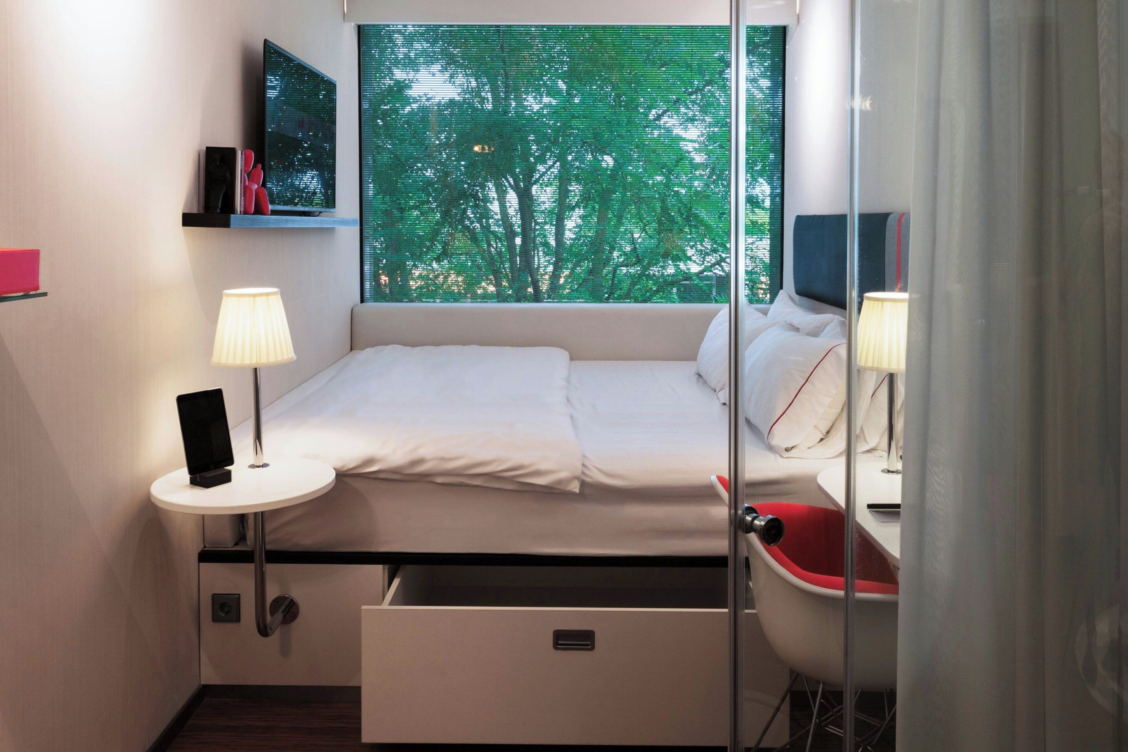 Ein kleines Hotelzimmer mit Bett, Nachttisch, Lampe und einem Fenster mit Blick auf Bäume.