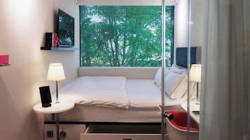 Ein kleines Hotelzimmer mit Bett, Nachttisch, Lampe und einem Fenster mit Blick auf Bäume.