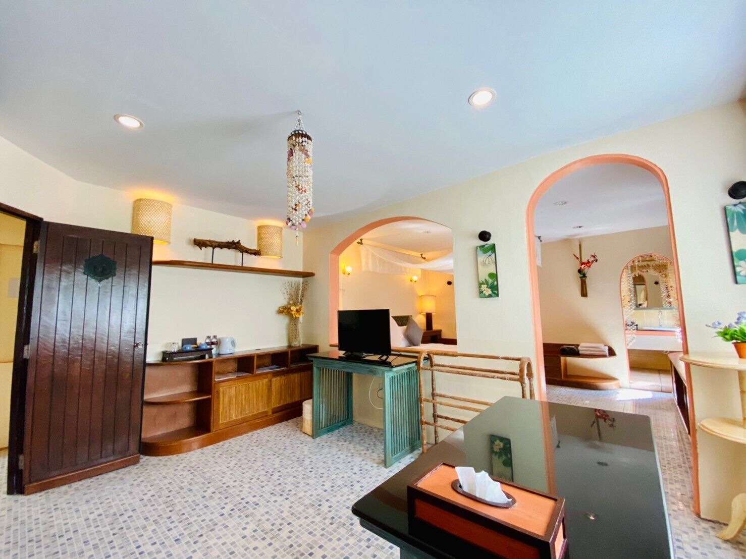 Une chambre d’hôtel avec une porte en bois, une télévision, une petite table avec une boîte de mouchoirs, un miroir et une salle de bain visible à travers une arche.