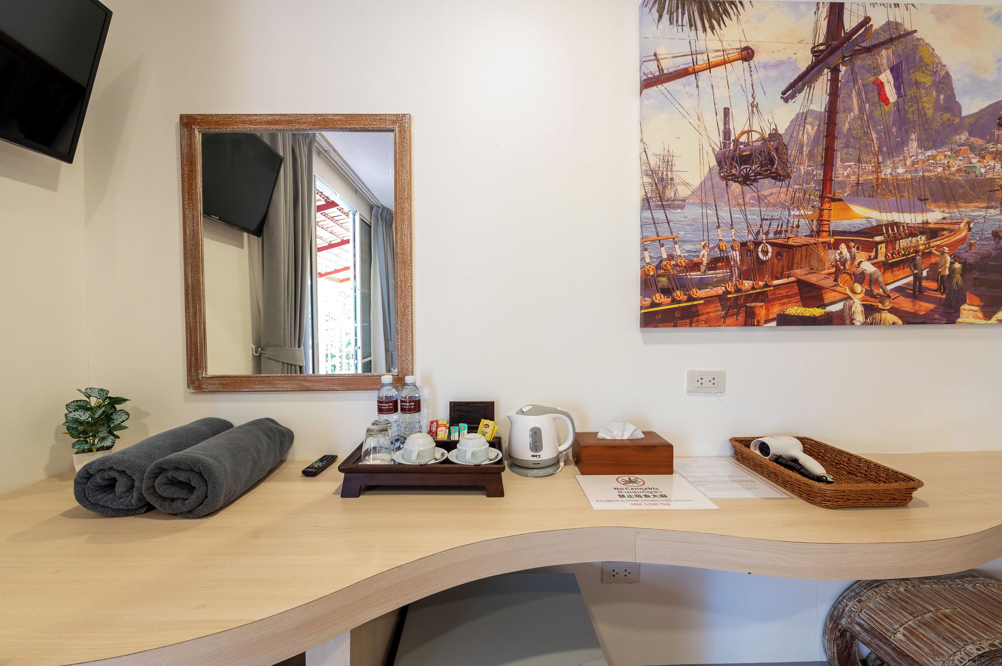 A hotel room with a wooden desk, a mirror, a painting of a harbor scene, a remote control, a tray with toiletries, and a basket with a hairdryer.
