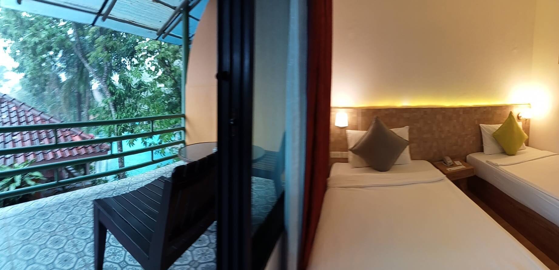 Una camera d'albergo con due letti, un balcone che si affaccia su alberi e un edificio con tetto rosso, e un tavolino.