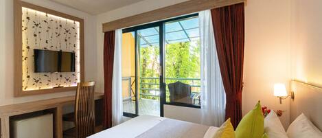 Habitación de hotel con cama, televisor montado en la pared, escritorio con silla y balcón con vistas a zonas verdes.