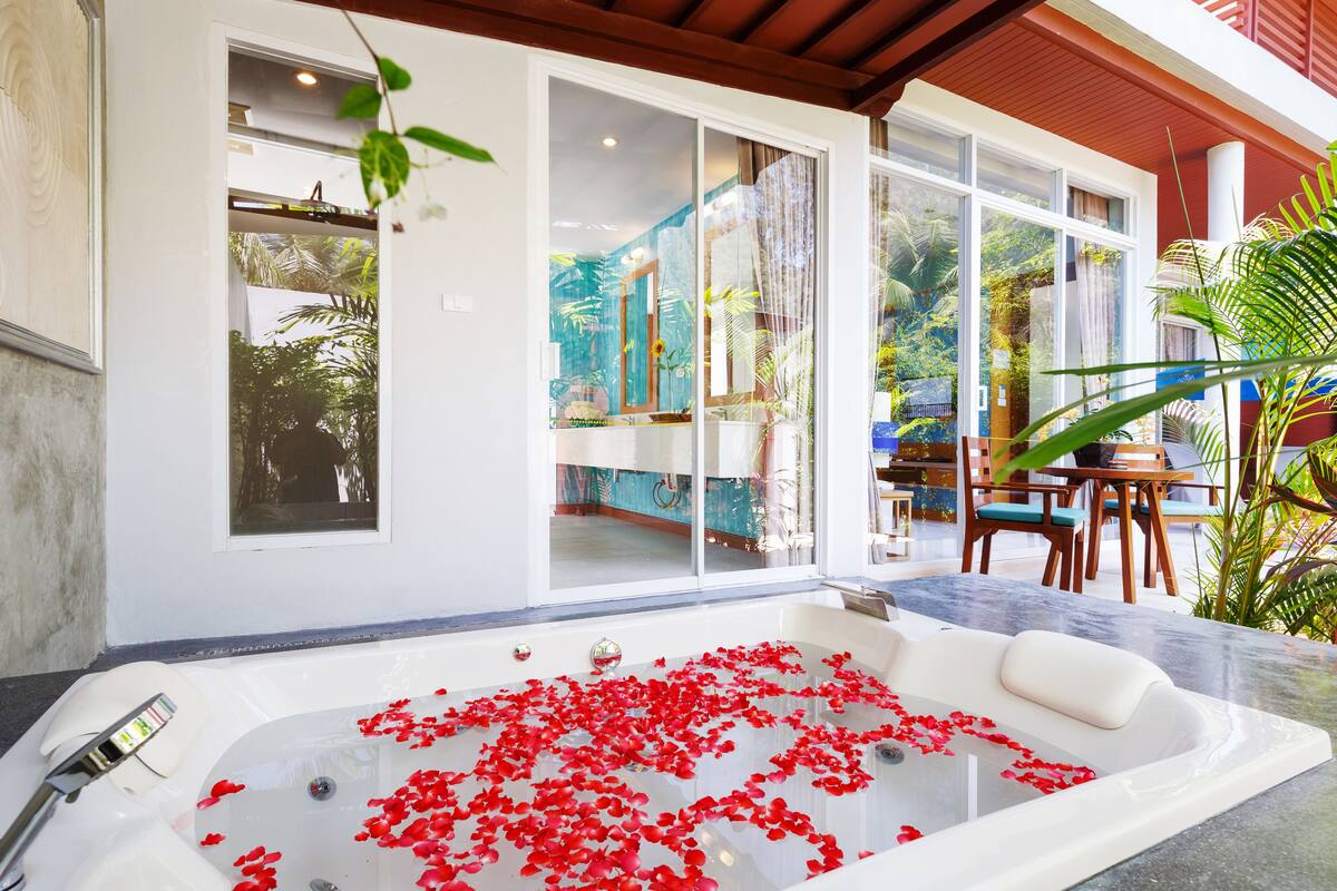 A spa area with a large bathtub filled with water and red rose petals, a wooden deck, and a view of the surrounding greenery.