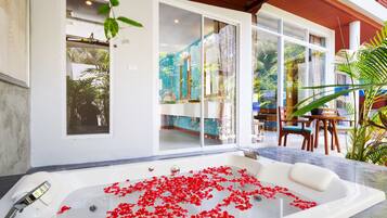 Un área de spa con una bañera grande llena de agua y pétalos de rosa roja, una terraza de madera y vistas al entorno verde.