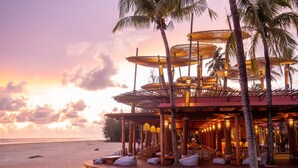 Un resort frente a la playa con zonas de estar al aire libre, palmeras y vistas al atardecer.