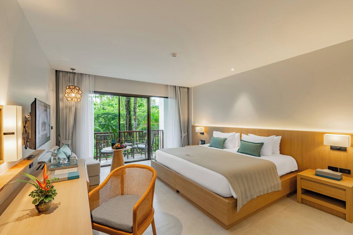 A modern hotel room with a large bed, a desk with a computer, a chair, a balcony with a table and chairs, and a view of greenery outside.