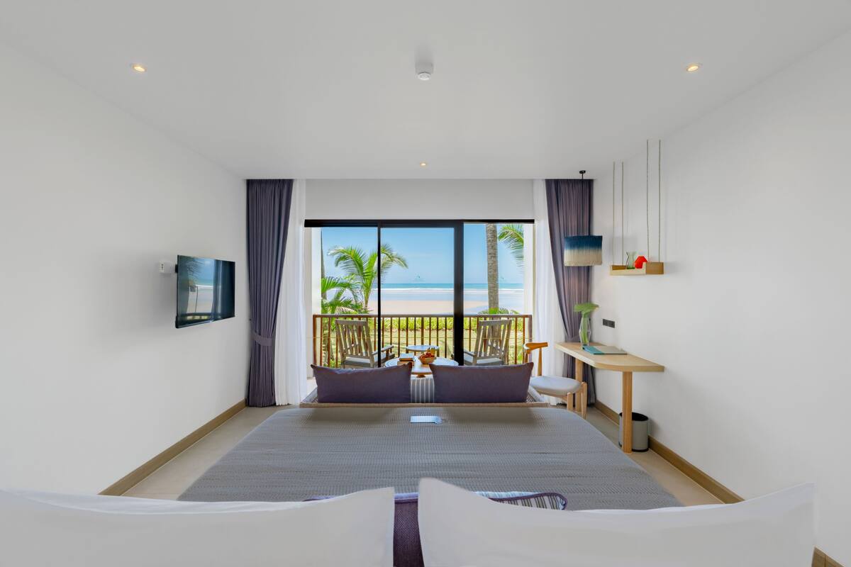 A modern hotel room with a large bed, a desk, and a view of the beach.