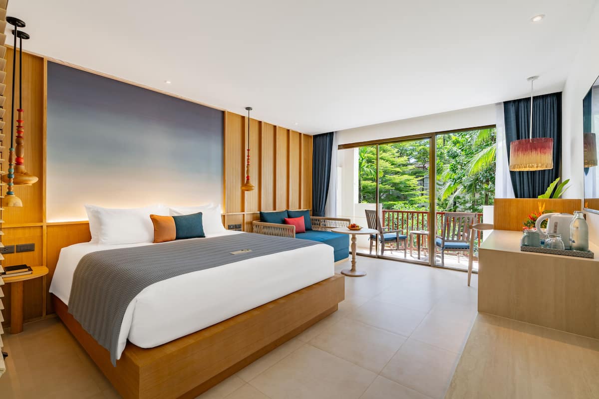 A modern hotel room with a large bed, a balcony with a view of greenery, a sitting area with a table, and a reception desk.
