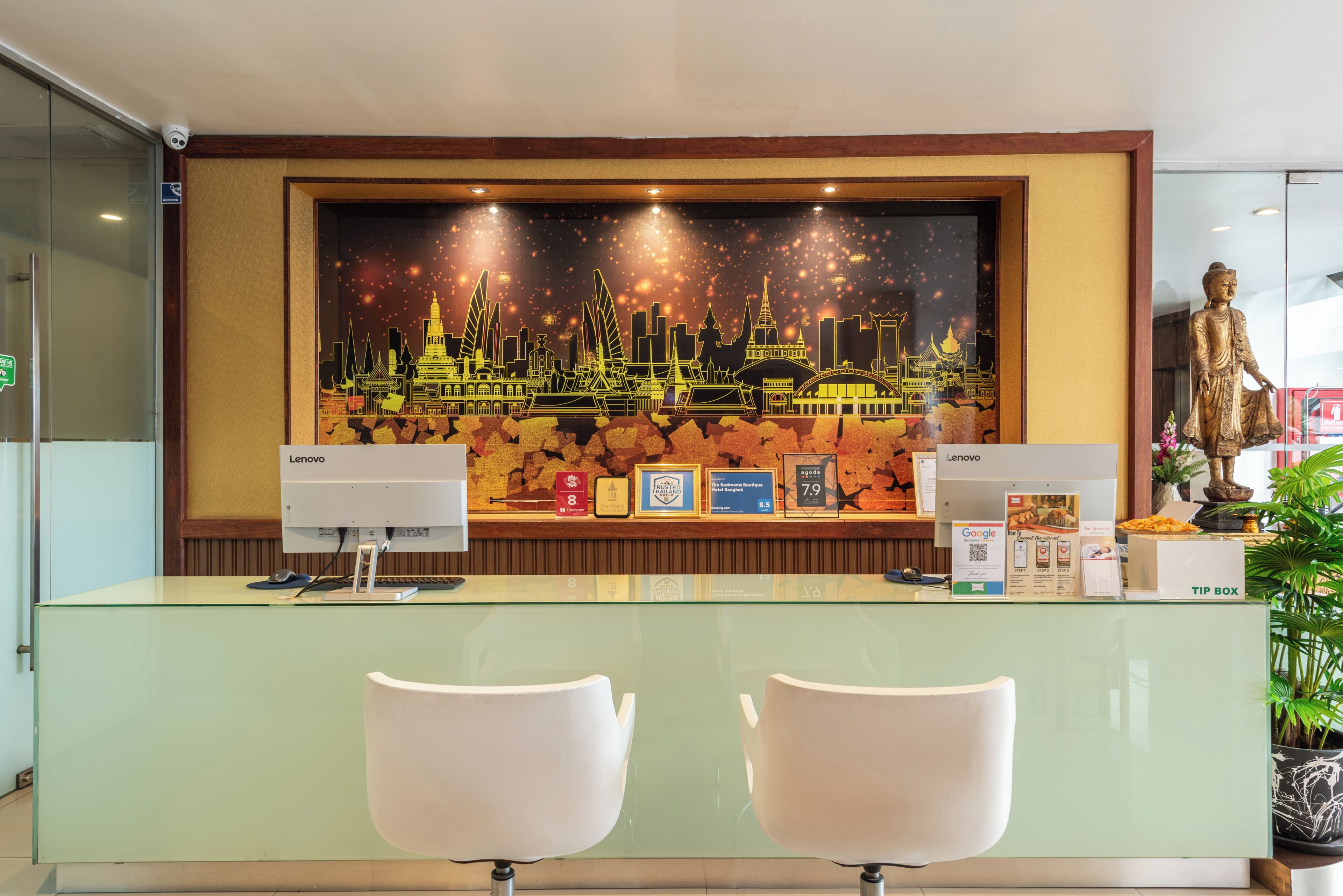 Una recepción con un mural de la ciudad, dos sillas blancas, una computadora Lenovo, un bote para propinas y una estatua.