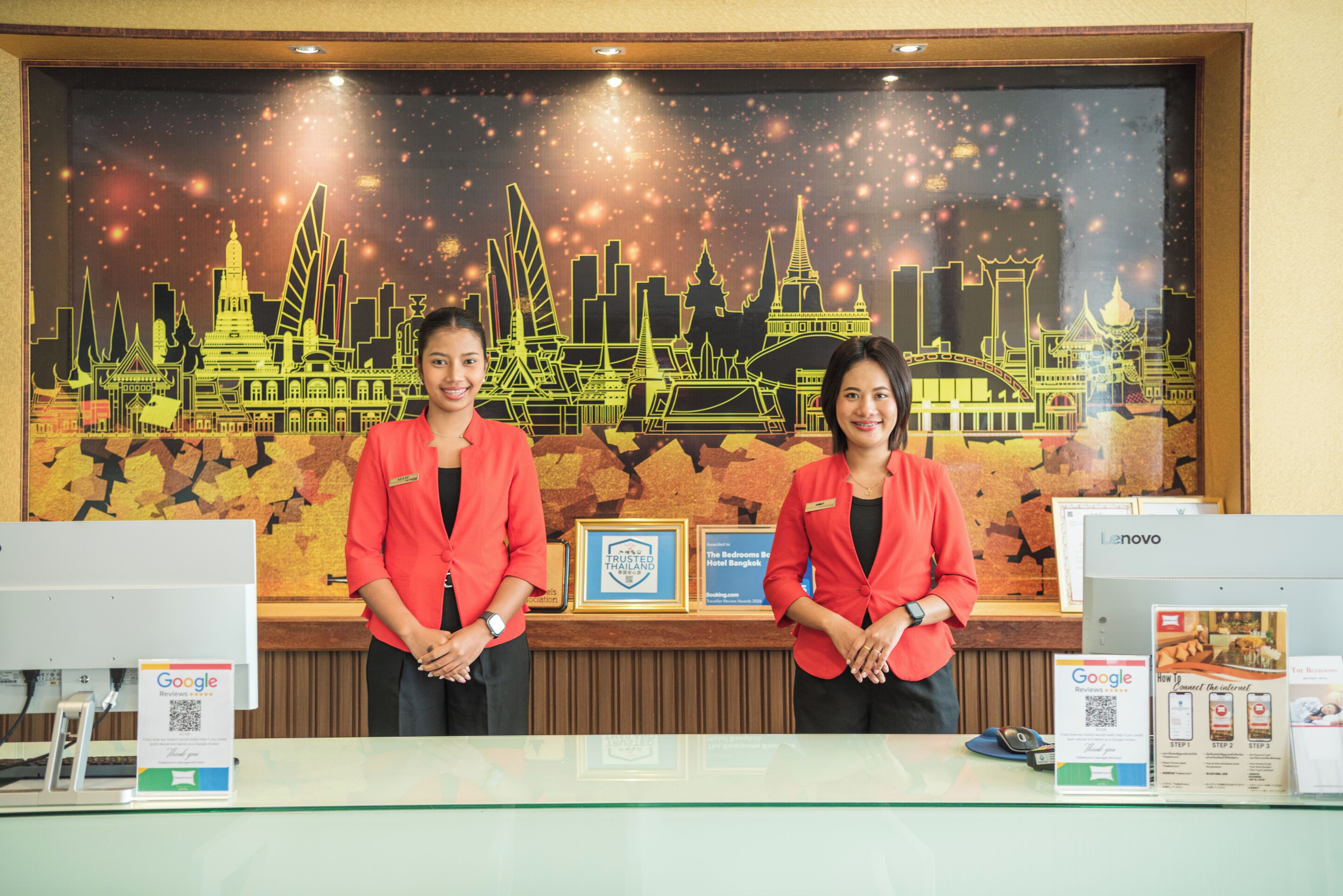 Two receptionists at a hotel desk with a cityscape mural, framed awards, and a Lenovo computer.