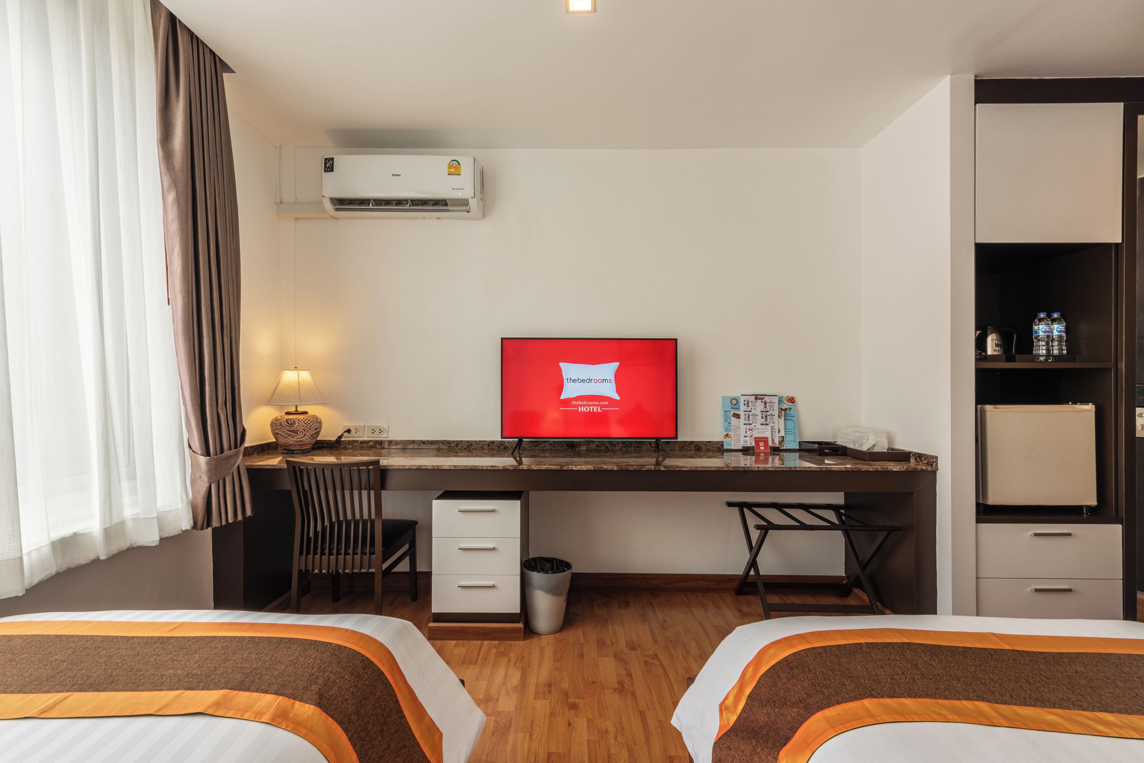 A hotel room with two beds, a desk, a chair, a TV, an air conditioner, a trash bin, and a minibar.