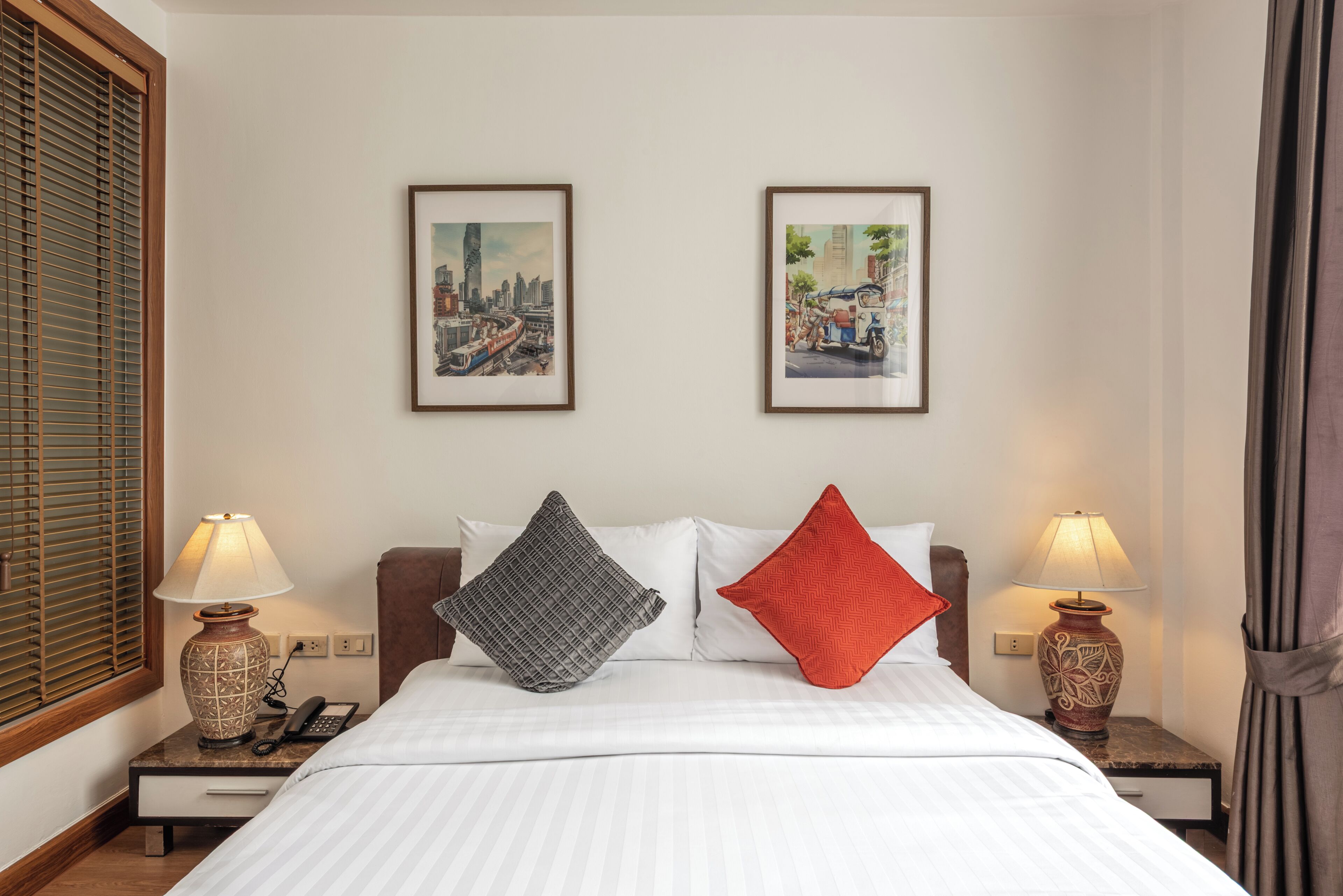 Hotelkamer met een bed, twee nachtkastjes met lampen, een telefoon, twee ingelijste foto's aan de muur en een raam met jaloezieën.