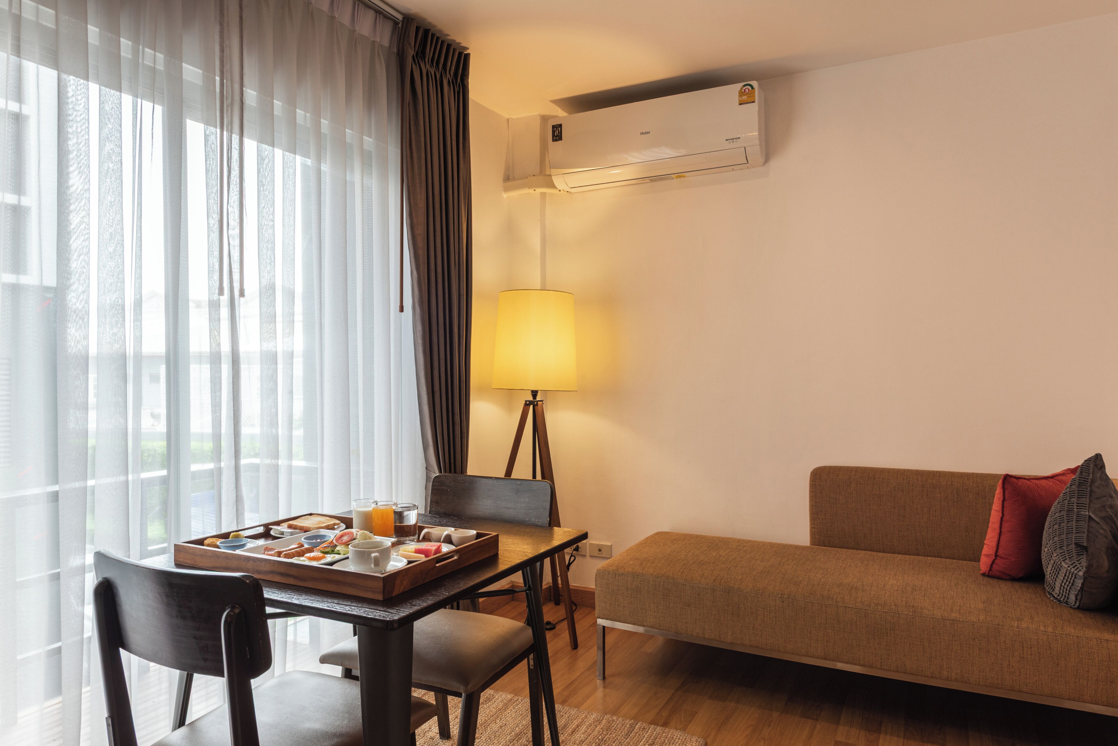A hotel room with a dining table, a sofa, a lamp, and an air conditioning unit.