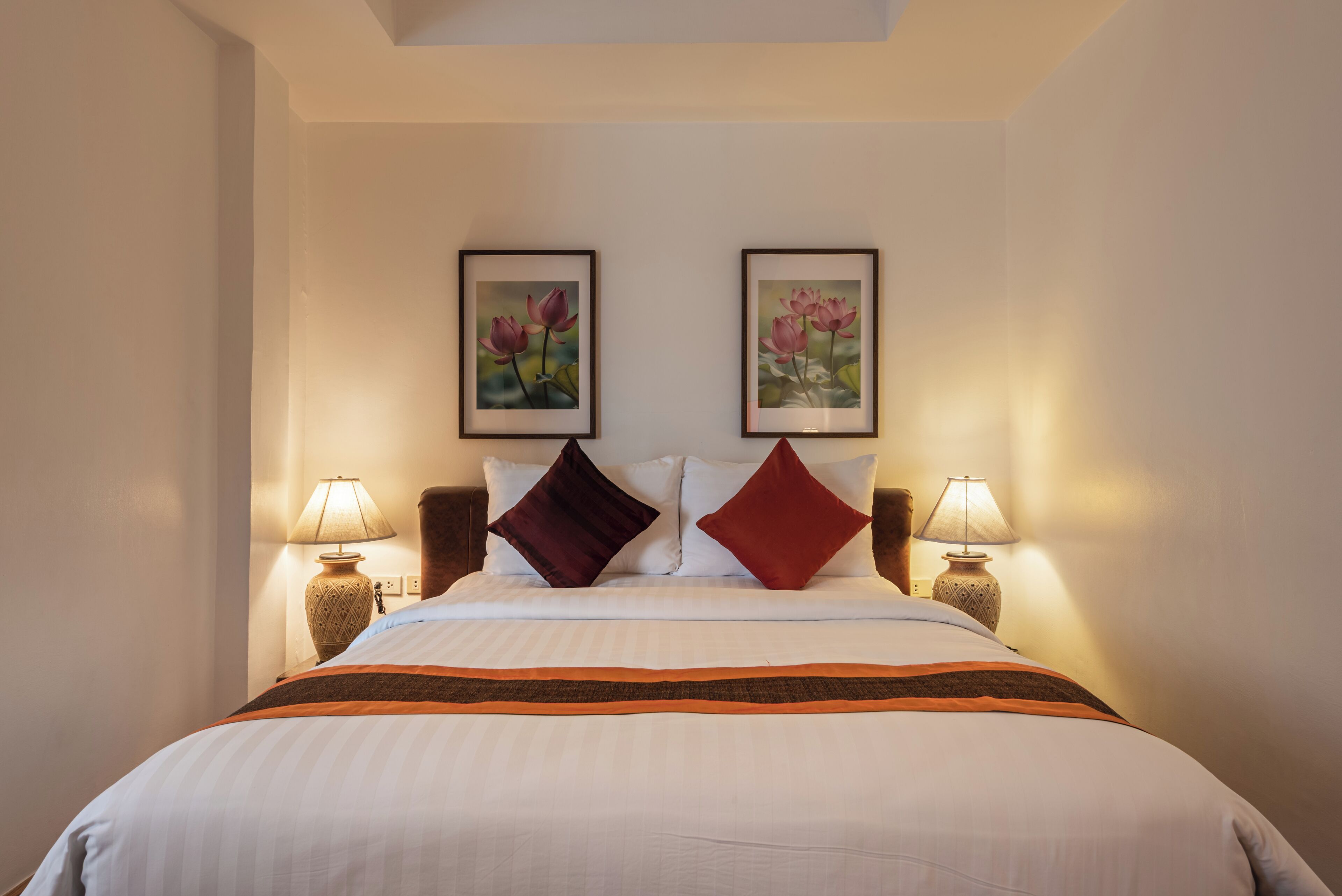 A hotel room with a bed, two framed floral paintings, two lamps, and a nightstand.