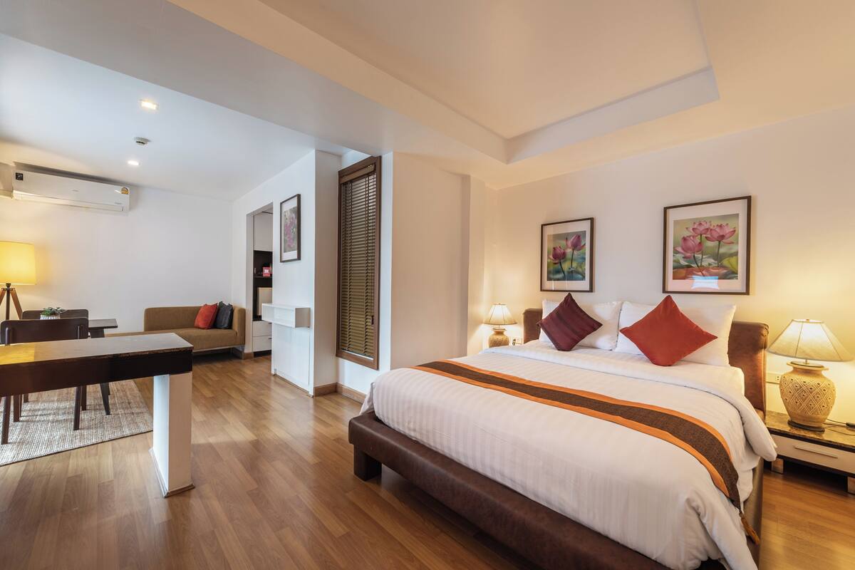 A hotel room with a bed, bedside tables, a desk, a sofa, and an air conditioning unit.