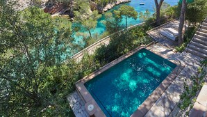 A pool surrounded by trees, overlooking a coastal area with buildings and boats. - Hotel Petit Cala Fornells (Calvia)