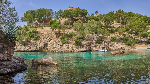 A coastal area with clear turquoise water, rocky terrain, and buildings on a hillside. - Hotel Petit Cala Fornells (Calvia)