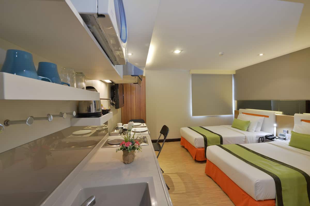 A hotel room with a kitchenette, two beds with green and white bedding, a small dining table, and a window.