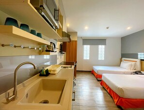 A hotel room with a kitchen area, a sink, a bed with orange bedding, and a window with patterned blinds. - Arawana Express Phromphong (Bangkok)