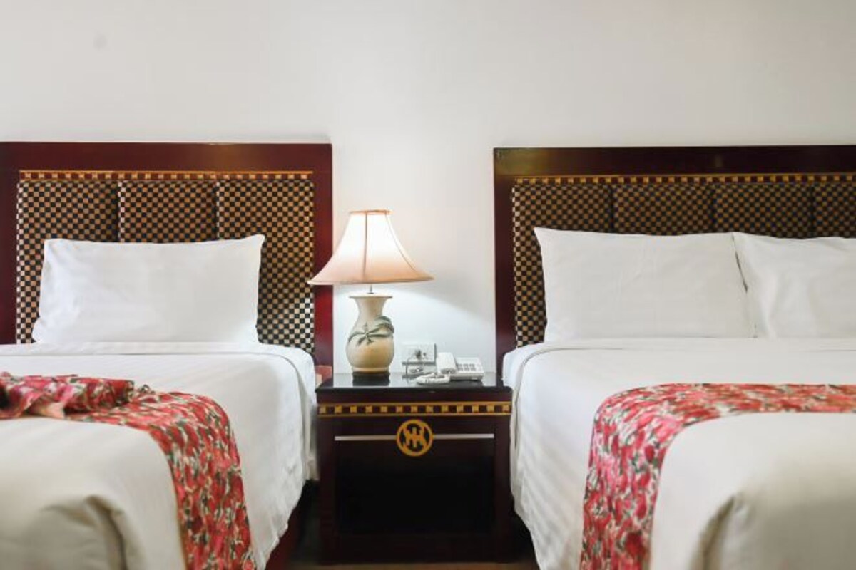 A hotel room with two beds, each with a checkered headboard, a bedside table with a lamp, and a small decorative vase.