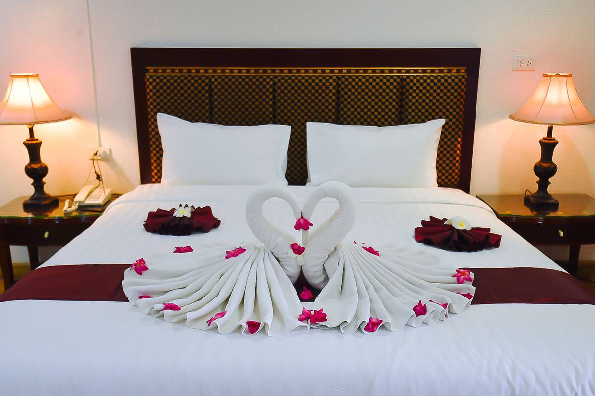 A neatly made bed with a heart-shaped towel arrangement and rose petals, two bedside lamps, and a checkered headboard.