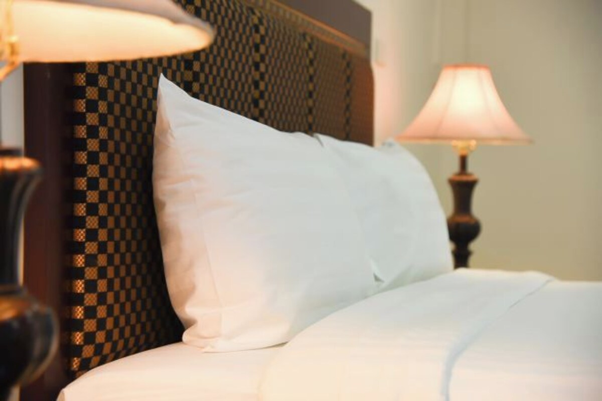A neatly made bed with white bedding and a single white pillow, a patterned wall, and a bedside lamp.