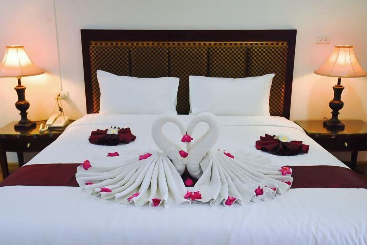 Un letto rifatto con cura, un asciugamano disposto a forma di cuore e petali di rosa, due lampade da comodino e un telefono sul tavolino.