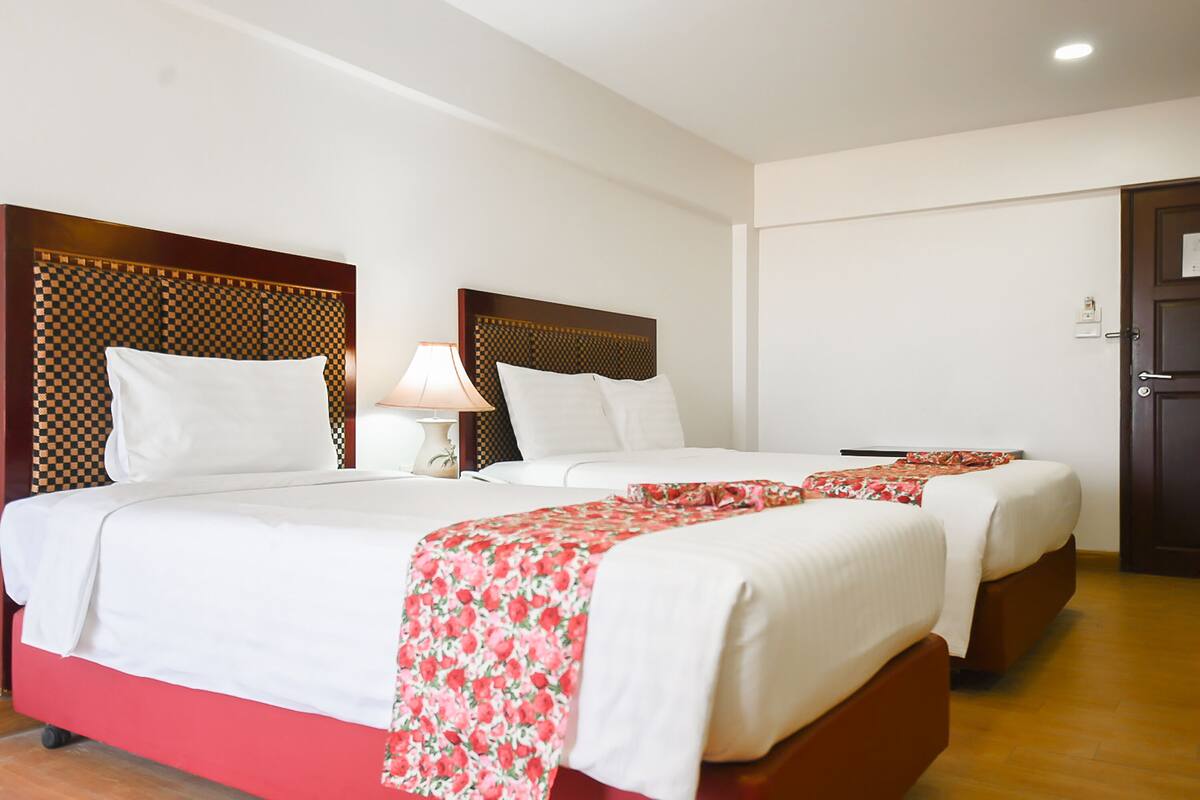 A hotel room with two beds, each with a wooden headboard, white bedding, and a red floral throw.