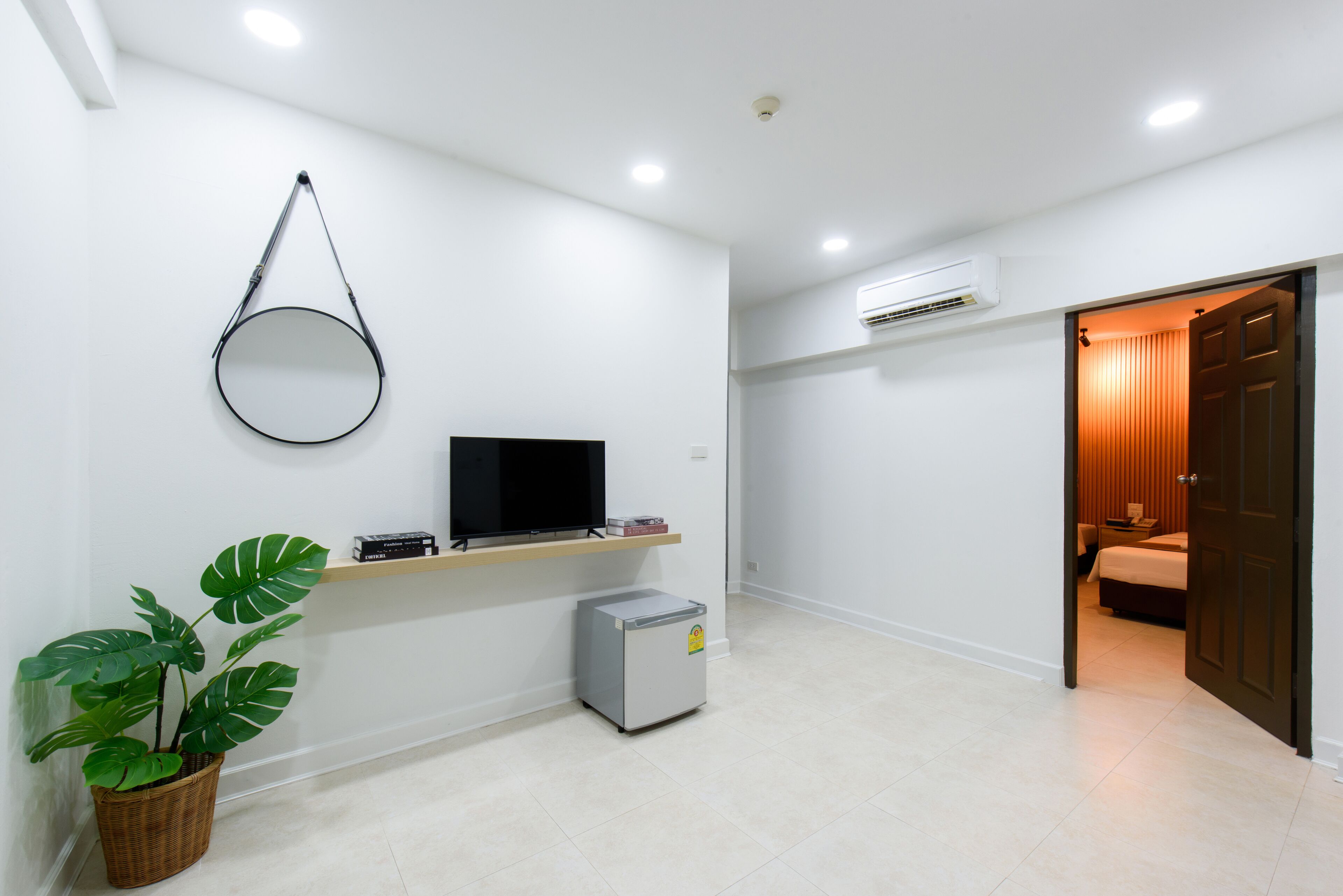 A hotel room with a bed, a TV, a small refrigerator, a plant, and a door leading to another room.