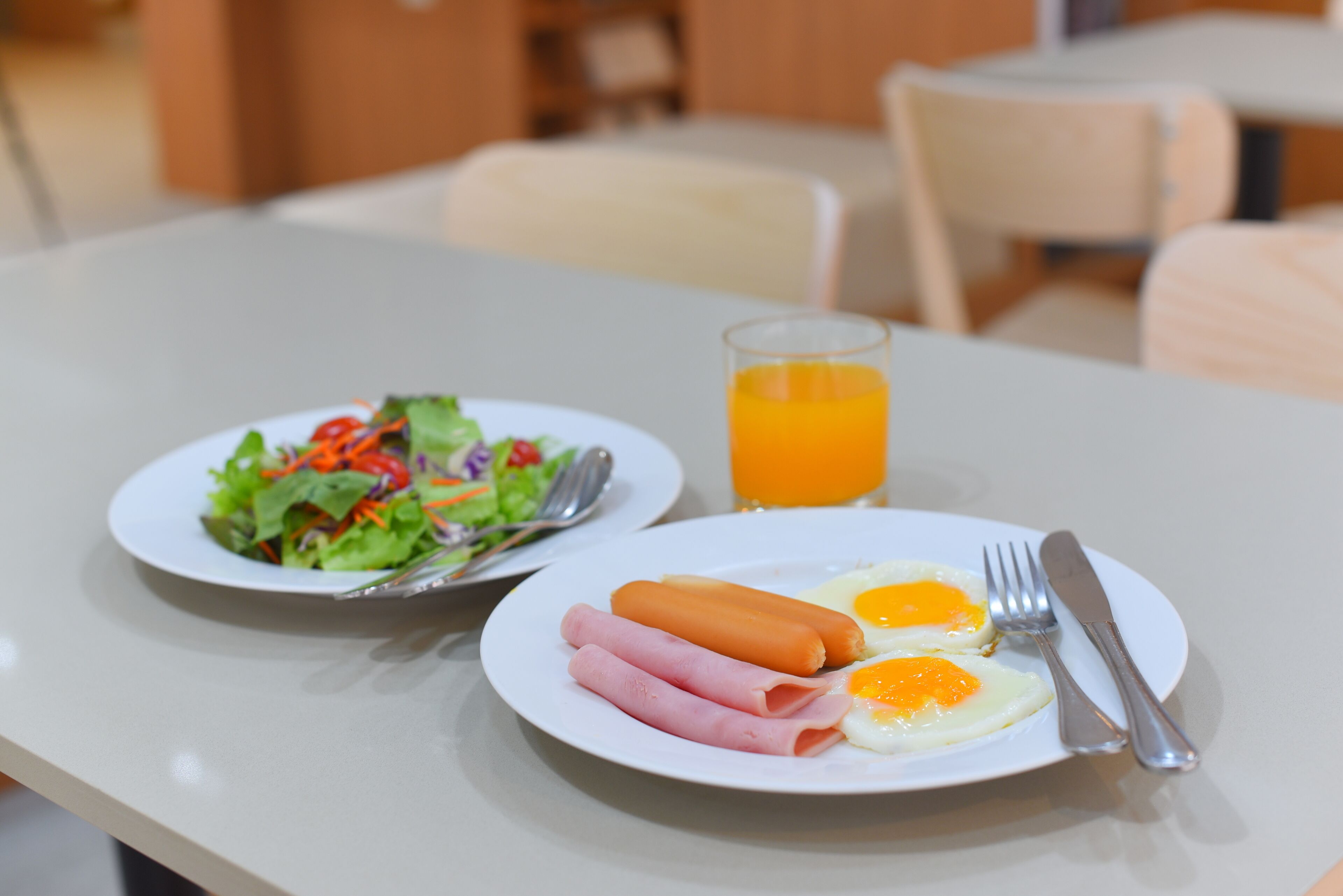A breakfast setting with a plate of eggs, ham, and a hot dog, and a plate of salad with a glass of orange juice.