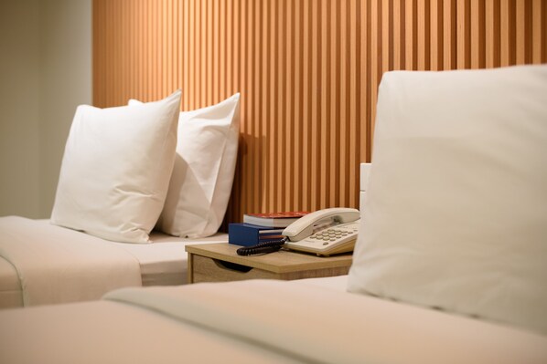 Una cama bien hecha con almohadas blancas y una mesita de noche con un teléfono, libros y un control remoto.