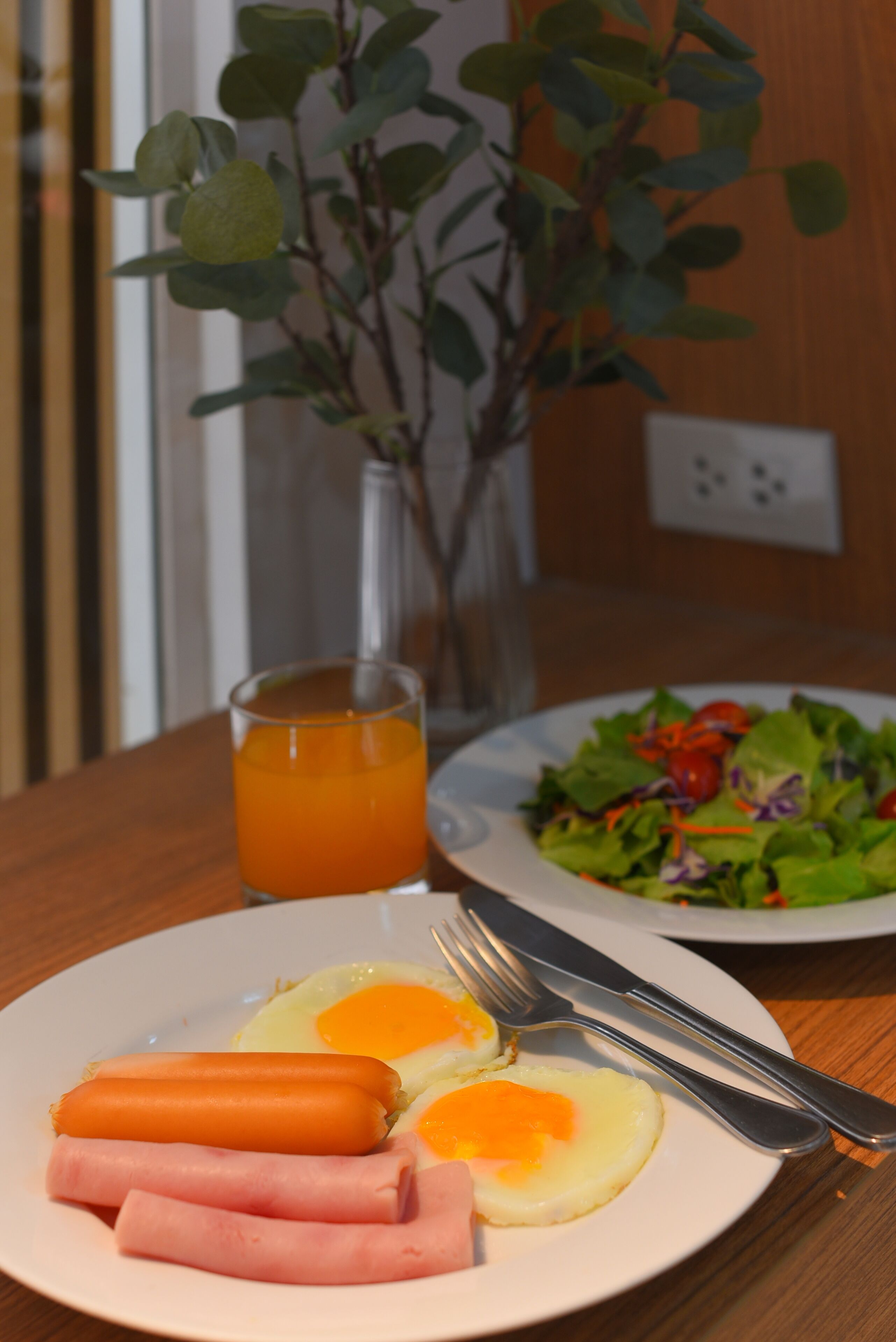 Un plato con huevos, jamón y salchichas, un vaso de zumo de naranja y una ensalada sobre una mesa de madera.