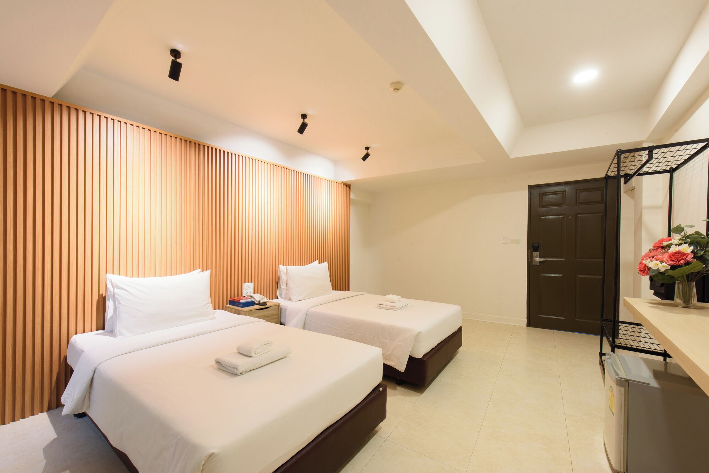 Habitación de hotel con dos camas, cabecera de madera con listones, mesita de noche, escritorio con silla, refrigerador y puerta con marco negro.