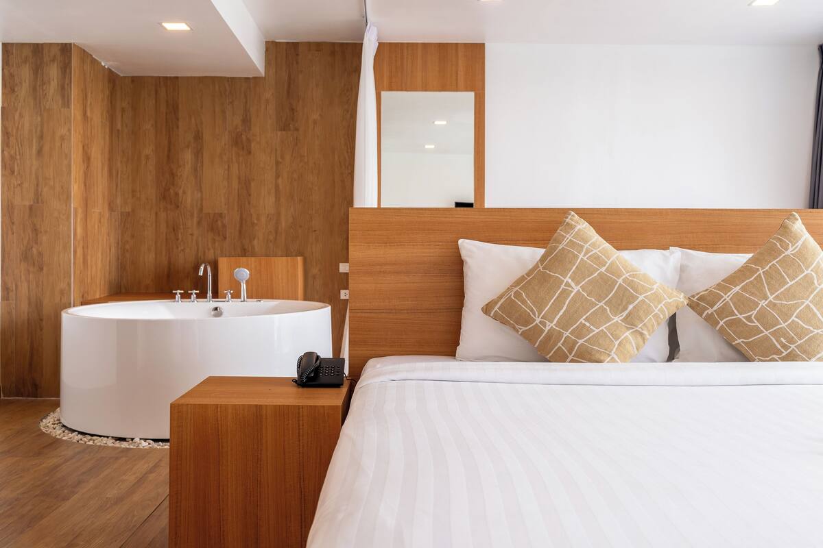 A modern hotel room with a wooden headboard, a large bed with white bedding, two decorative pillows, a round white bathtub, and a bedside table with a phone.