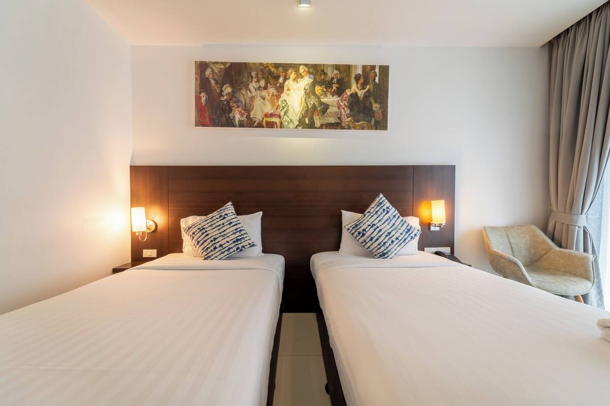 A hotel room with two single beds, a large painting above the beds, a chair, and a nightstand with a lamp.