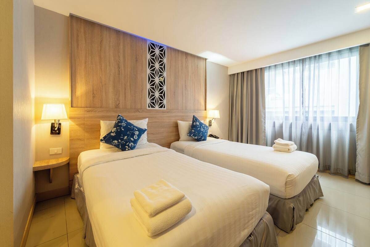 A hotel room with two beds, a wooden headboard, a decorative panel, and a window with curtains.