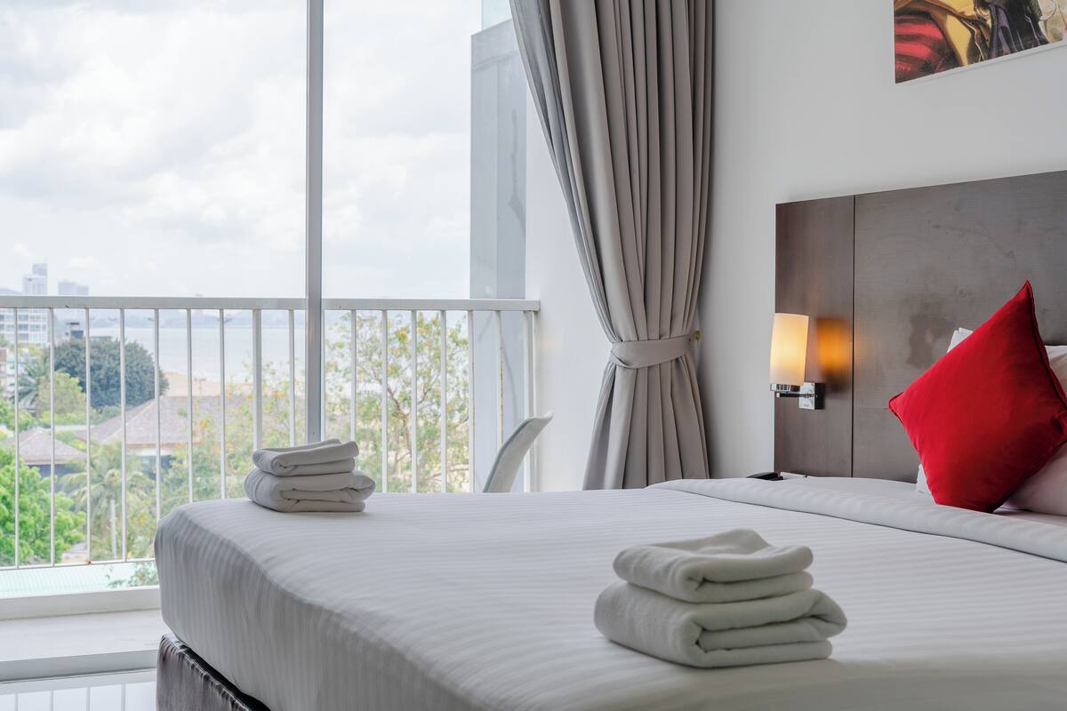 A hotel room with a large window, a bed with white linens, a red pillow, and a neatly folded towel on the bed.