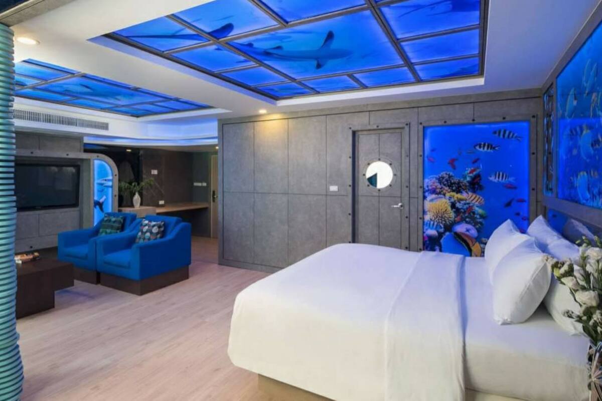 A modern hotel room with a large bed, a seating area with blue sofas, a wall aquarium, and a skylight with a blue ocean design.
