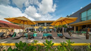 A pool area with sun loungers and umbrellas, a stone wall with plants, and a building with large windows in the background. - Kamala Beachfront Apartment (Kamala)